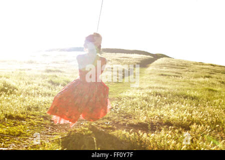 View from behind of a girl in a floral pink dress swinging on a rope swing with a path through a sunny field in the background. Stock Photo