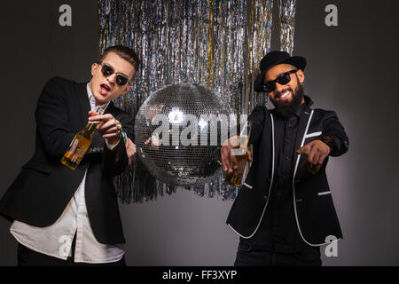 Two attractive smiling young men in suits and sunglasses with beer dancing and pointing on you over blck background Stock Photo