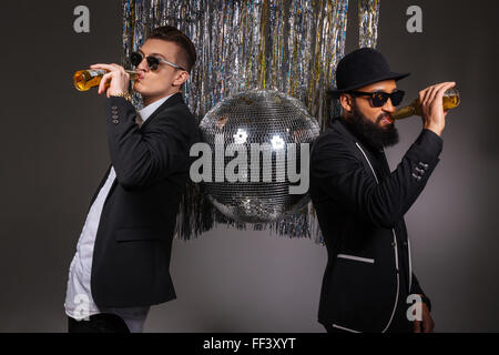 Two handsome confident young men in black suits and sunglasses standing near disco ball and drinking beer over black background Stock Photo