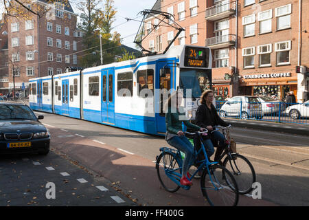 Two cyclists / bike / bikes / cycles and a Line 24 Dutch tram running through the centre of Amsterdam. Holland, The Netherlands. Stock Photo