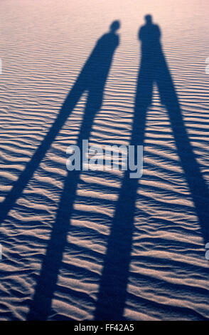 The setting sun casts the long shadows of two visitors walking across wind-rippled sand dunes near Stovepipe Wells in Death Valley National Park in California, USA. The geography of this vast park ranges from such desert sands to colorful badlands, rugged canyons, and snow-covered mountains. Recording some of the hottest temperatures in the world, Death Valley lives up to its forbidding name. Stock Photo