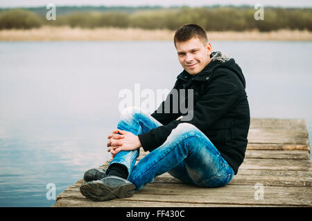 Young handsome man sitting on wooden pier in autumn day, relaxing,  thinking, smiling. Casual style - jeans, jacket Stock Photo