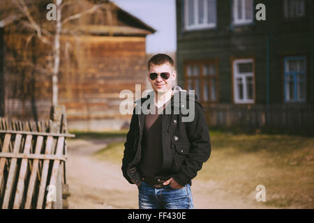 Young handsome man staying near old wooden house in autumn day, relaxing,  thinking, smiling. Casual style - jeans, jacket, sung Stock Photo