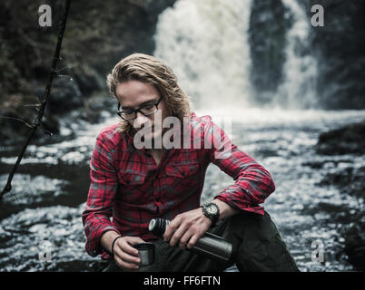 A young man sitting pouring a hot drink from a flask by a fast flowing stream. Stock Photo