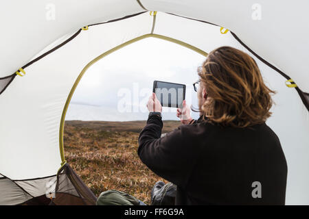 A man sitting in the shelter of a tent looking out, taking a photograph with a digital tablet Stock Photo