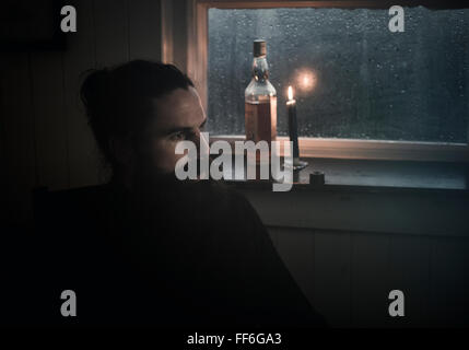 A man sitting in the dark by a window in candlelight drinking from a small glass.  A bottle beside him. Stock Photo