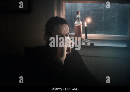 A man sitting in the dark by a window in candlelight drinking from a small glass.  A bottle beside him. Stock Photo