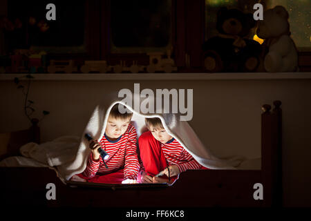 Two sweet boys, reading a book in bed after bedtime, using flashlights, winter night Stock Photo