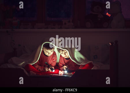 Two sweet boys, reading a book in bed after bedtime, using flashlights, winter night Stock Photo