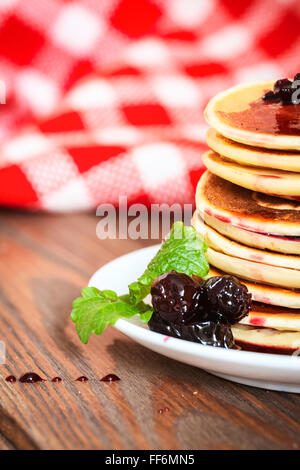 stack of pancakes on a white plate with cherry jam, mint leaves, red checkered napkin, brown wooden background of boards, rustic Stock Photo