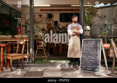 Portrait of a young man standing in front of his coffee shop. Young man with beard wearing an apron  standing with her arms cros Stock Photo