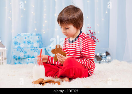 Cute little happy boy, eating cookies and drinking milk, waiting for Santa in pajama on Christmas Eve night Stock Photo