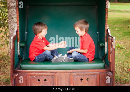Two kids, brothers,sitting in a sheltered bench, playing hand clapping game, outdoors in the summer Stock Photo