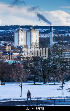 A man walks through the park with two blocks of flats, a smoking chimney and a wind turbine on Glasgow South side taken from Queens Park, Glasgow. Stock Photo