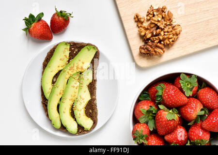 high-angle shot of an avocado toast in a white plate, a white bowl full of strawberries and some peeled walnuts on a wooden chop Stock Photo
