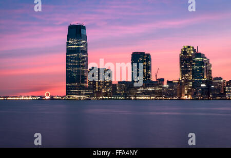 A beautiful sunset over the skyscrapers of Paulus Hook Jersey City and the Hudson River, with a pink sky reflected in the water.