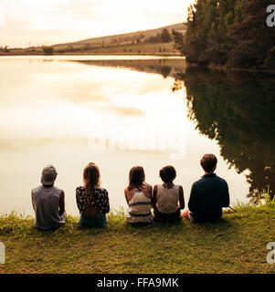 Rear view image of group of young friends sitting in a row by a lake and looking at a beautiful landscape view. Group of friends Stock Photo