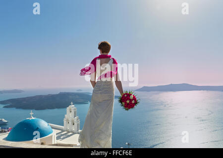 Bride with a bouquet of flowers in his hands stands high over the coast. Santorini, Greece. Stock Photo