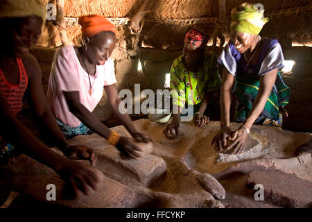 A group of women in Fada N’Gourma Burkina Faso grind organic millet on a traditional table carved from stone to make flour. The table has been in the village for years and has well molded troughs to collect the flour. Stock Photo