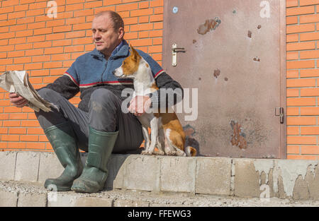 Young dog and mature man reading crumpled newspaper while sitting on a threshold at warm spring day Stock Photo