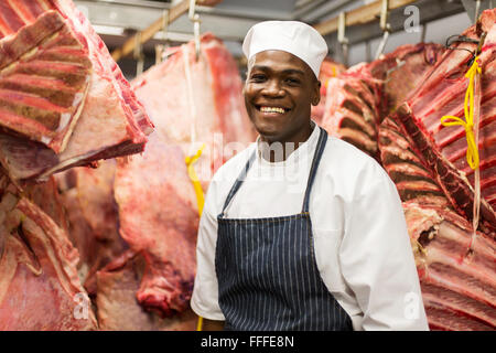 young African male butcher standing against meat hanging in butchery Stock Photo