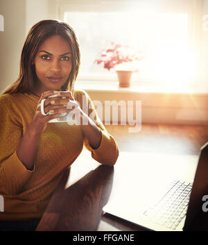 https://l450v.alamy.com/450v/ffgane/beautiful-black-woman-in-yellow-sweater-sitting-at-table-holding-coffee-cup-next-to-laptop-with-bright-light-coming-through-wind-ffgane.jpg