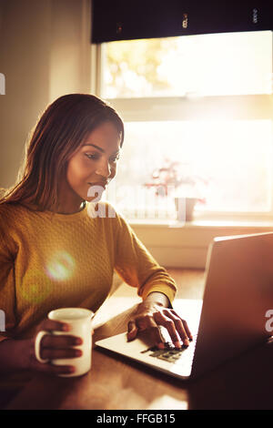 Happy gorgeous adult single female sitting near bright sunny window typing on her laptop computer and holding coffee cup Stock Photo