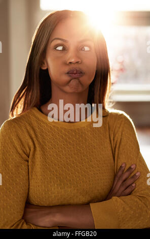 Young woman making a goofy face squinting and puffing out her cheeks in exasperation as she stands with folded arms in front of Stock Photo