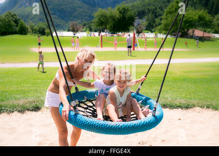 Blonde mom pushes the girls on the round rope swing in a playground. Stock Photo