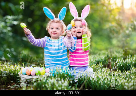 Kids on Easter egg hunt in blooming spring garden. Children with bunny ears searching for colorful eggs in snow drop flower Stock Photo