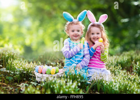 Kids on Easter egg hunt in blooming spring garden. Children with bunny ears searching for colorful eggs in snow drop flower Stock Photo
