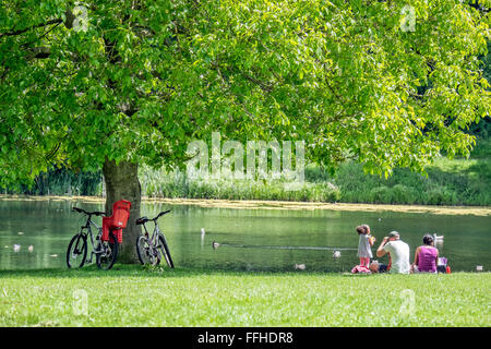 A family relaxing under the shade of a tree, by a lake with their bicycles rested against the tree trunk Stock Photo