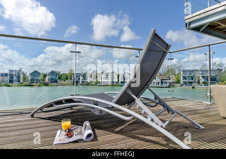 A sun lounger on a decking, waterside veranda overlooking a lake & waterfront homes, South Cerney, Cotswolds, UK Stock Photo