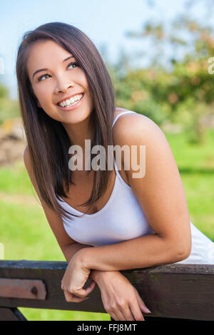 Beautiful happy Asian Eurasian young woman or girl wearing white t-shirt, smiling and leaning on fence in sunshine Stock Photo