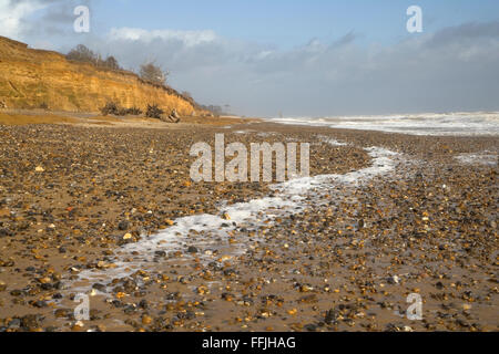 Beach at Covehithe consisiting of sand and pebbles, with spume on beach caused by stormy sea, with crumbling cliffs in the backg Stock Photo