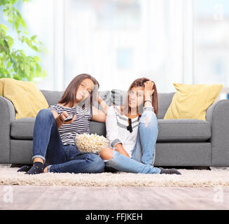 Two bored teenage girls watching TV seated on the floor in front of a gray sofa at home shot with tilt and shift lens Stock Photo