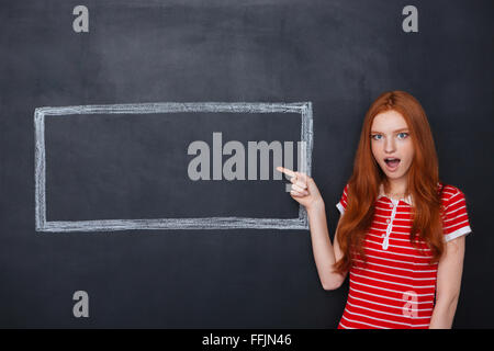 Amazed cute redhead woman pointing on copyspace inside frame drawn on blackboard background Stock Photo