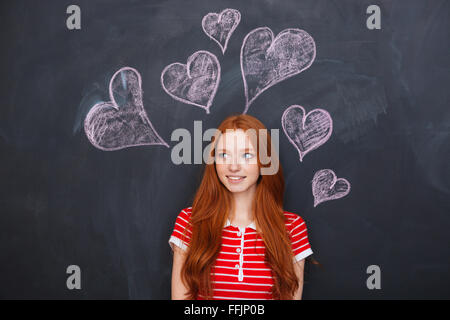 Beautiful tender young woman with long red hair standing over chalkboard with drawn hearts behind her Stock Photo