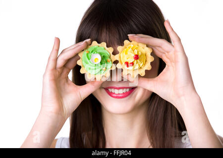 Headshot portrait of young smiling woman holding two cream tart cakes in dough baskets, fooling around, using biscuits as eyes Stock Photo