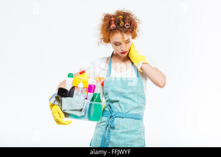 Exhausted redhead young woman in curlers and yellow gloves holding plastic box with cleaning supplies and having neck pain over  Stock Photo