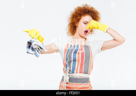 Shocked unhappy redhead young woman in striped apron closing nose by hand and holding dirty rag over white background Stock Photo