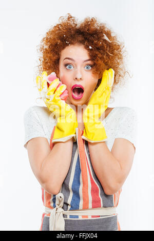 Amazed attractive young woman with foam on her curly red hair in protective gloves holding yellow sponge over white background Stock Photo