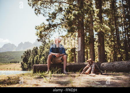 Portrait of a mature man sitting on a log staring out at the lake. Senior man camping by the lake on a summer day.