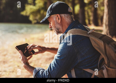Portrait of mature male hiker using digital tablet outdoors. Senior man with backpack sitting by a lake looking at his digital t Stock Photo
