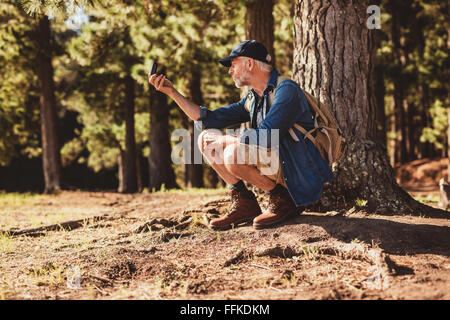 Mature man stops to check his position by using a compass while out on hike in the forest. male hiker sitting by a tree in fores Stock Photo