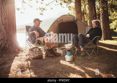 Portrait of happy senior couple sitting in chairs by tent at campsite. Mature man and woman relaxing and talking near a lake on Stock Photo