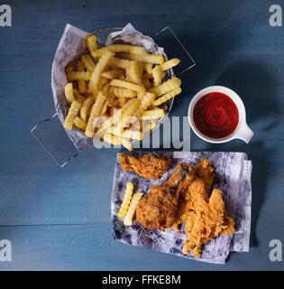 Chicken fries strips and legs on paper with basket of  French fries and bowl of ketchup sauce over blue wooden table. Top view. Stock Photo