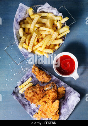 Chicken fries strips and legs on paper with basket of  French fries and bowl of ketchup sauce over blue wooden table. Top view Stock Photo