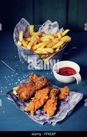 Chicken fries strips and legs on paper with basket of  French fries and bowl of ketchup sauce over blue wooden table. Turquoise Stock Photo