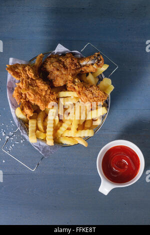 Chicken fries strips and legs with French fries in metal basket and bowl of ketchup sauce over blue wooden table with sea salt. Stock Photo